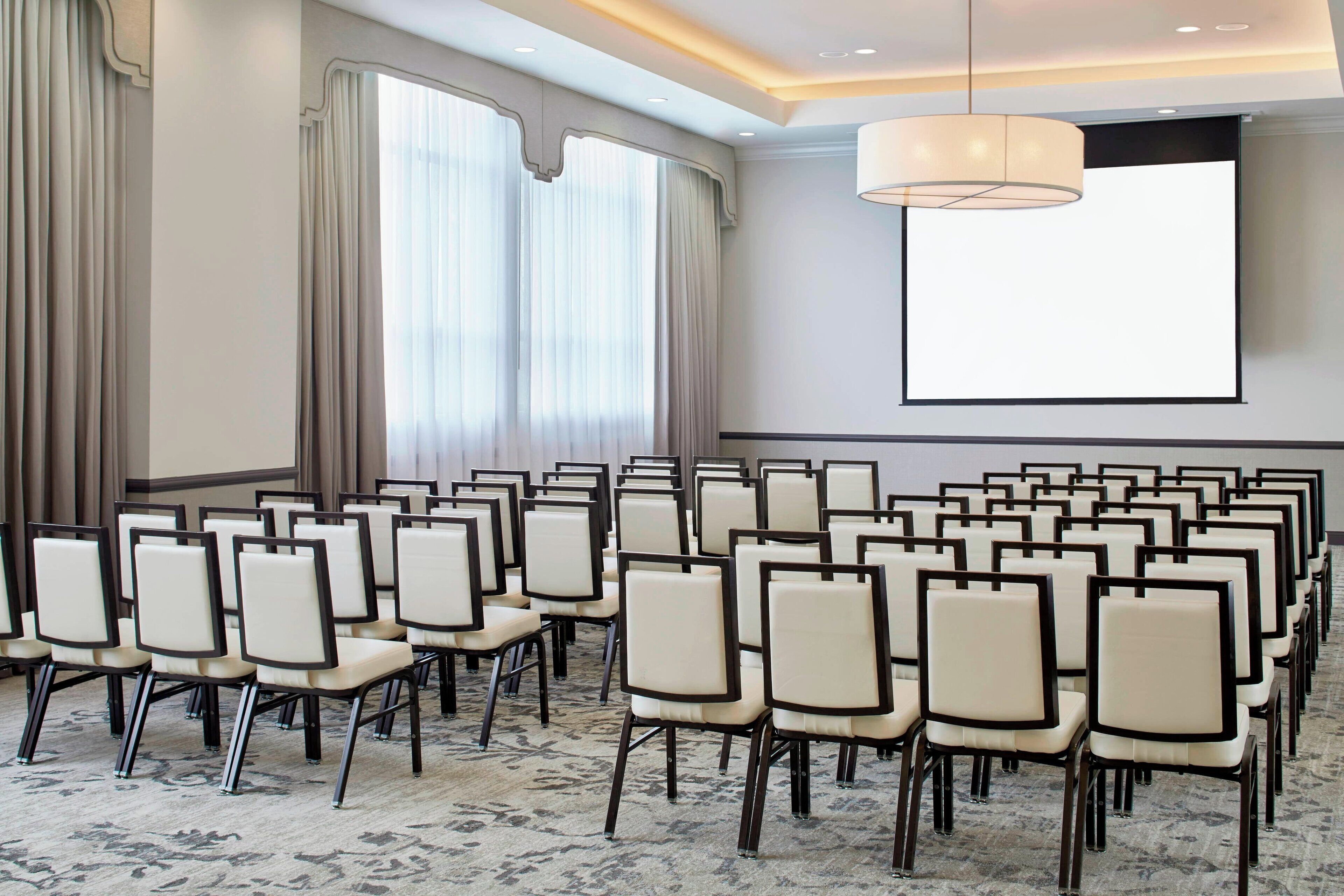 Ein Konferenzraum mit Reihen von Stühlen, einer Projektionsleinwand und großen Fenstern mit Vorhängen.