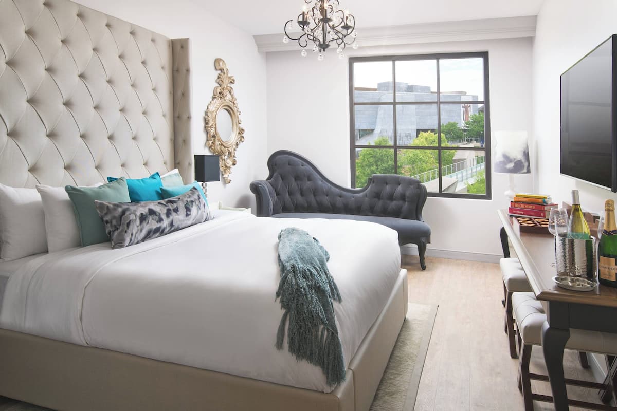 A bedroom with a large bed, a chaise lounge, a desk with a TV, and a view of a cityscape through a large window.