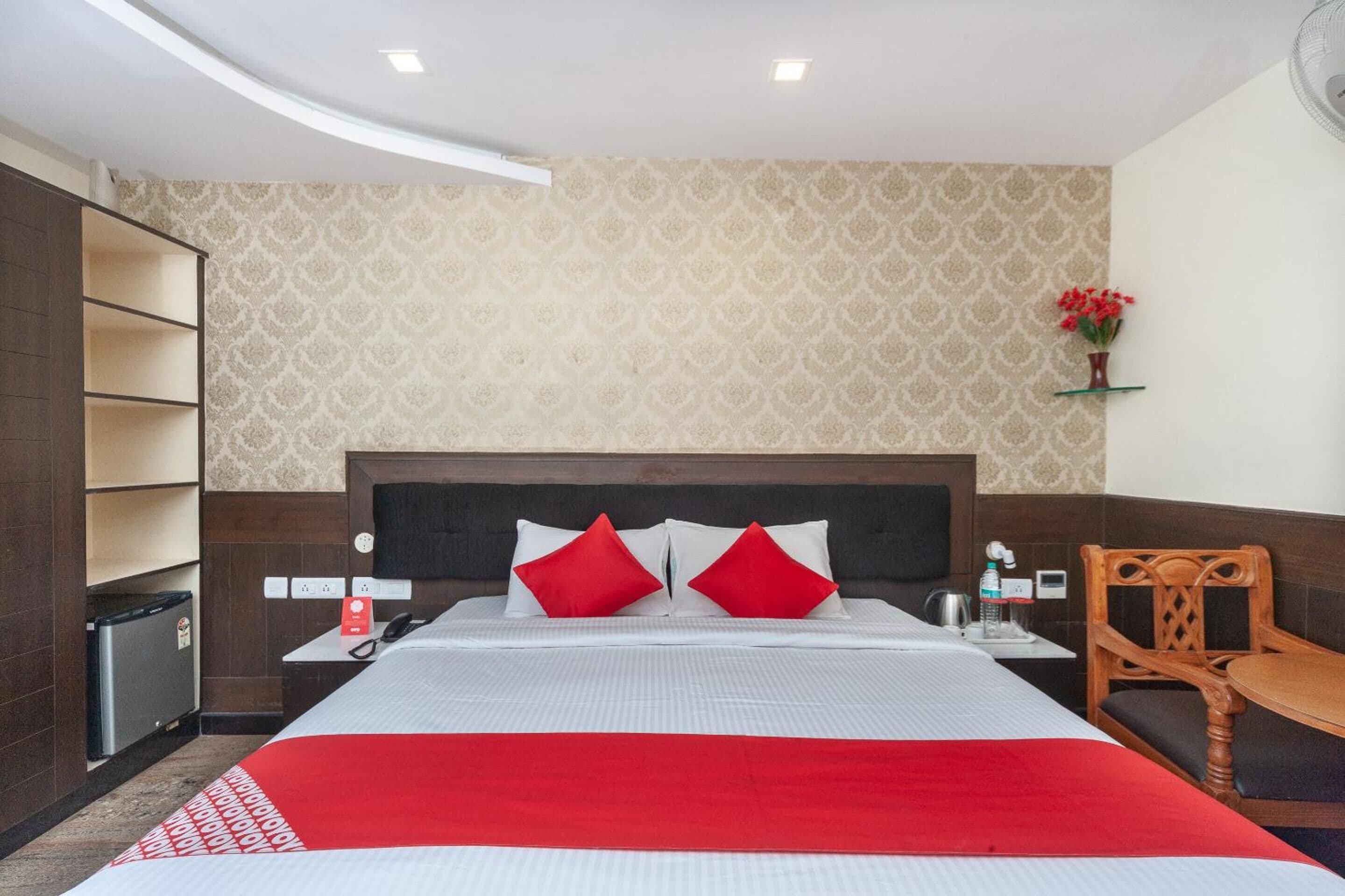 A hotel room with a bed, a wooden chair, a small table, a nightstand, a wall-mounted shelf with flowers, and a patterned wallpaper.