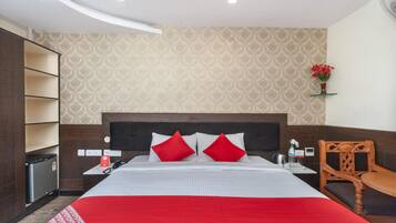 A hotel room with a bed, a wooden chair, a small table, a nightstand, a wall-mounted shelf with flowers, and a patterned wallpaper.