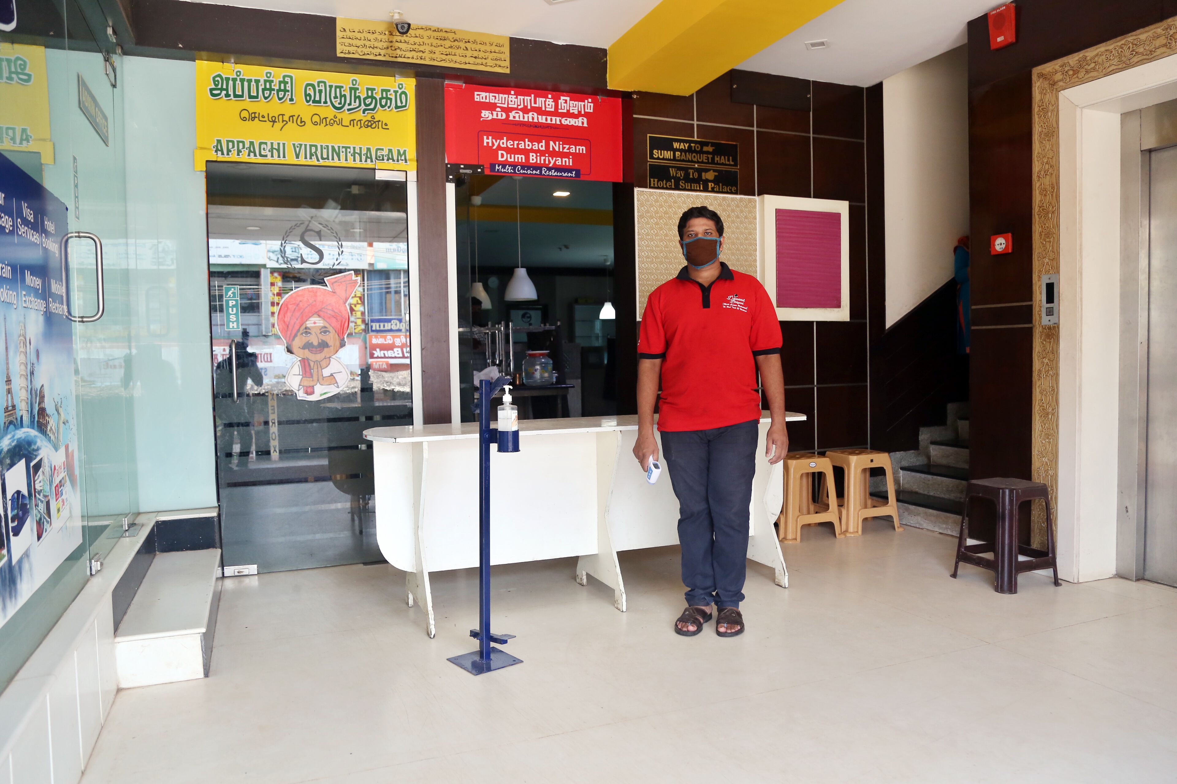 A man in a red shirt standing in a lobby with a reception desk, signs in multiple languages, and a staircase leading to an upper level.