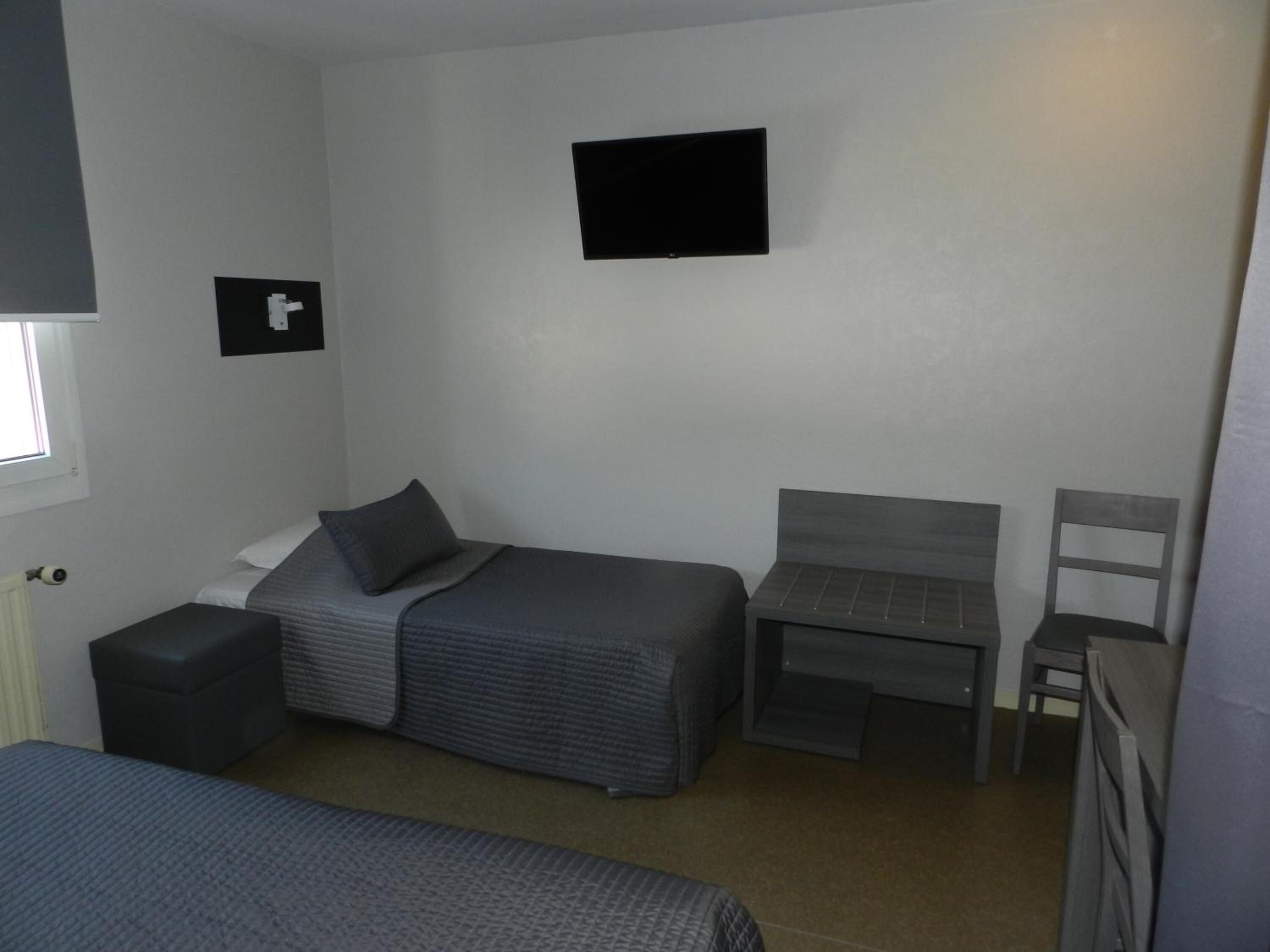 Una habitación pequeña y funcional con una cama individual, una mesita, una silla y un televisor fijado a la pared.