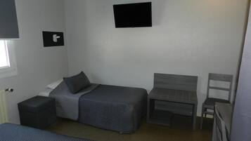 Una habitación pequeña y funcional con una cama individual, una mesita, una silla y un televisor fijado a la pared.