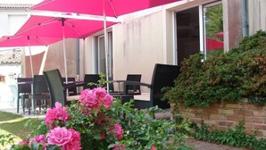 Un patio al aire libre con sombrillas rosas, sillas negras y un jardín con flores rosadas.
