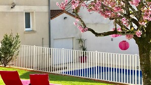 Zona de piscina con sillas reclinables rojas, una cerca blanca y un árbol con flores rosadas.