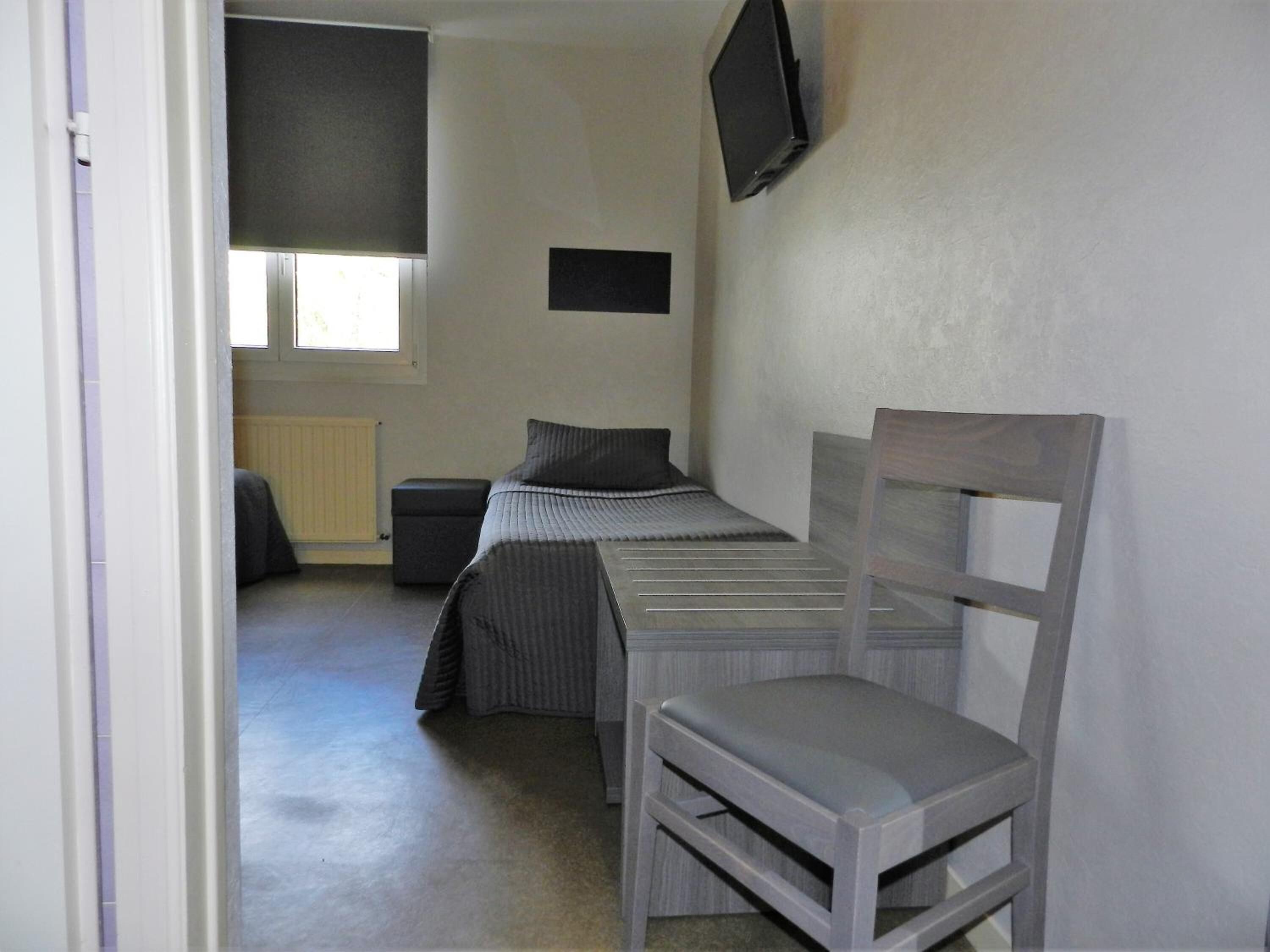 A small, sparsely furnished room with a single bed, a chair, a small table, a television mounted on the wall, and a window providing natural light.