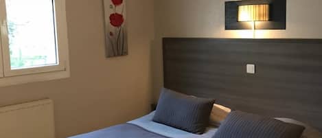 Une chambre à coucher comprenant un lit, une lampe fixée au mur, un radiateur et une fenêtre avec des stores.