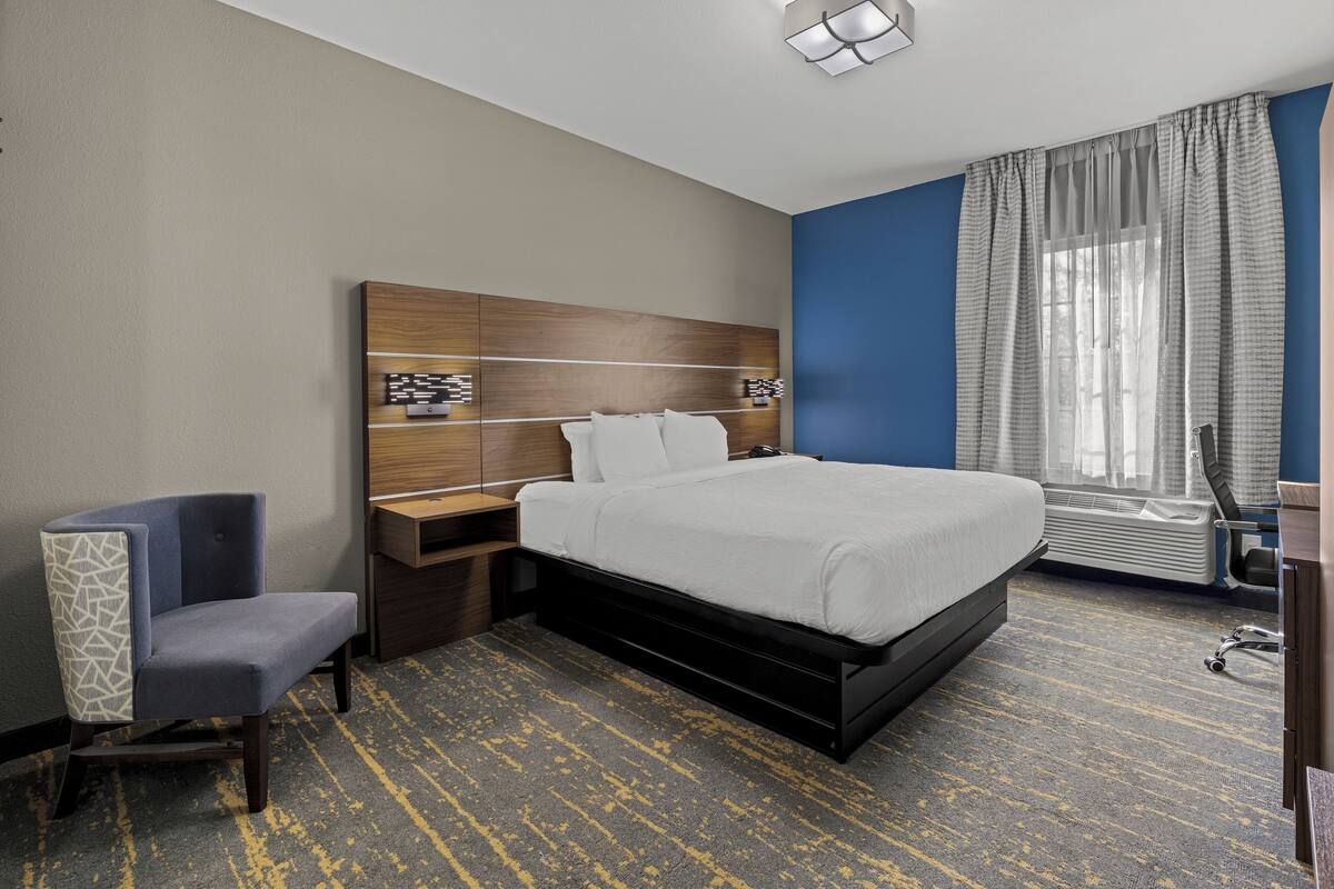 A hotel room with a bed, a chair, a nightstand, a desk, and a window with curtains.
