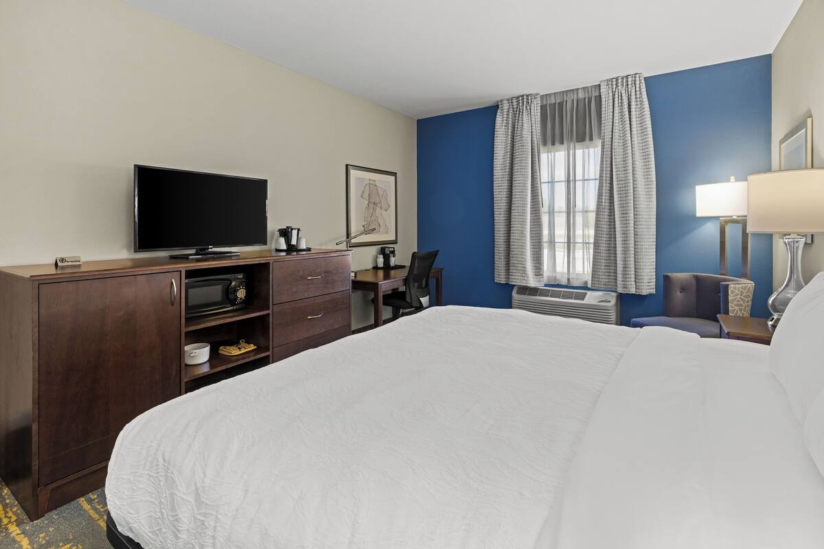 A hotel room with a large bed, a desk with a chair, a television, a desk lamp, a framed picture, and a window with curtains.