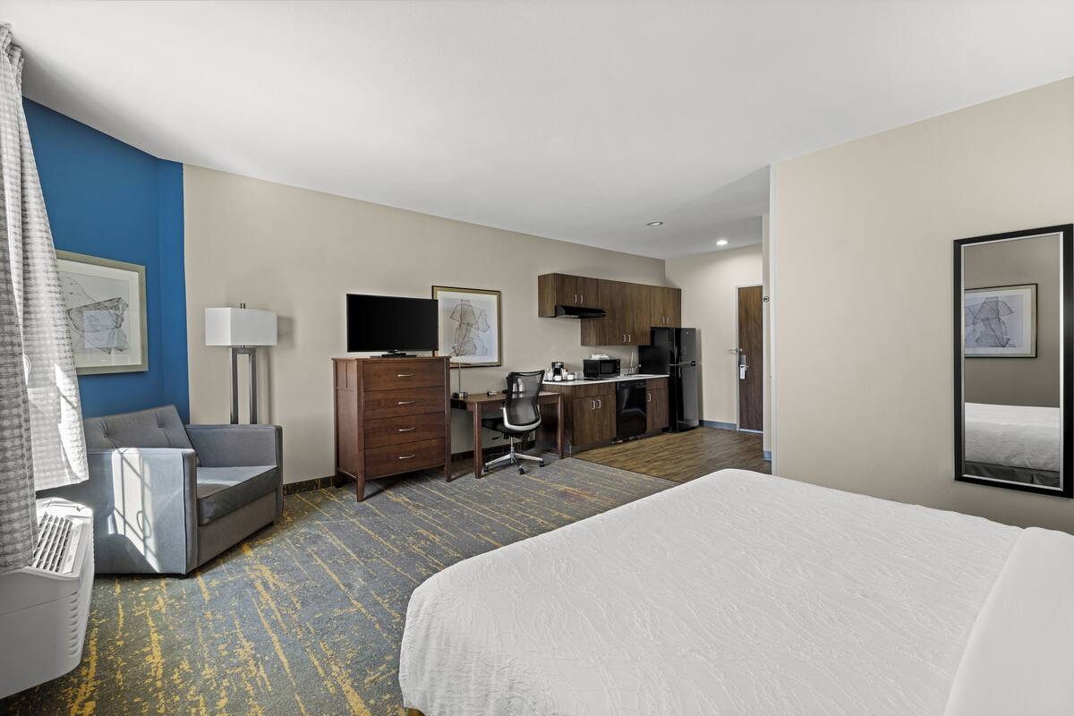 A hotel room with a bed, a desk, a chair, a television, a kitchen area, and artwork on the walls.