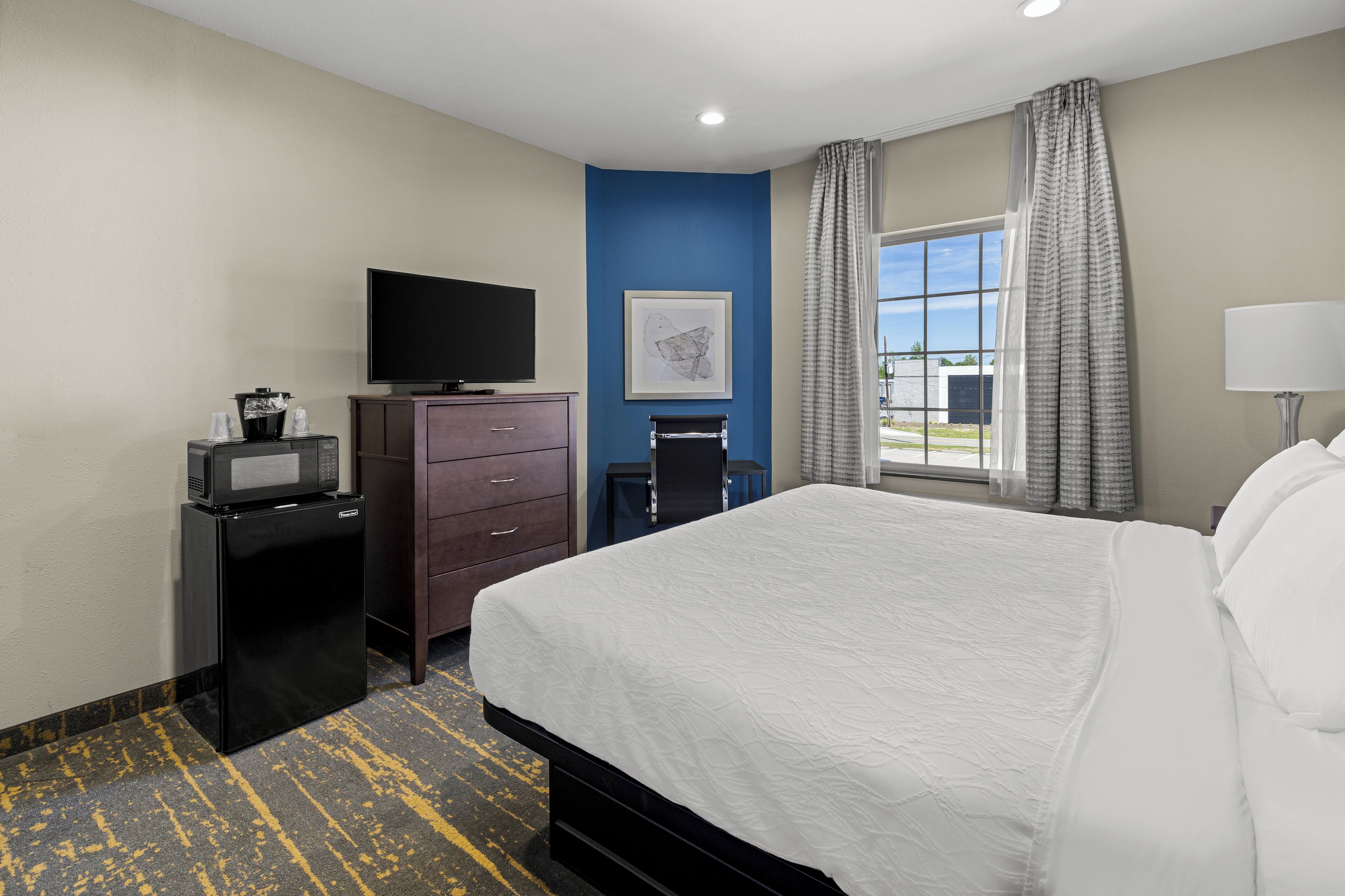 A hotel room with a bed, a television, a desk with a computer, a window with curtains, and a painting on the wall.