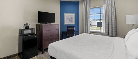 A hotel room with a bed, a television, a desk with a computer, a window with curtains, and a painting on the wall.