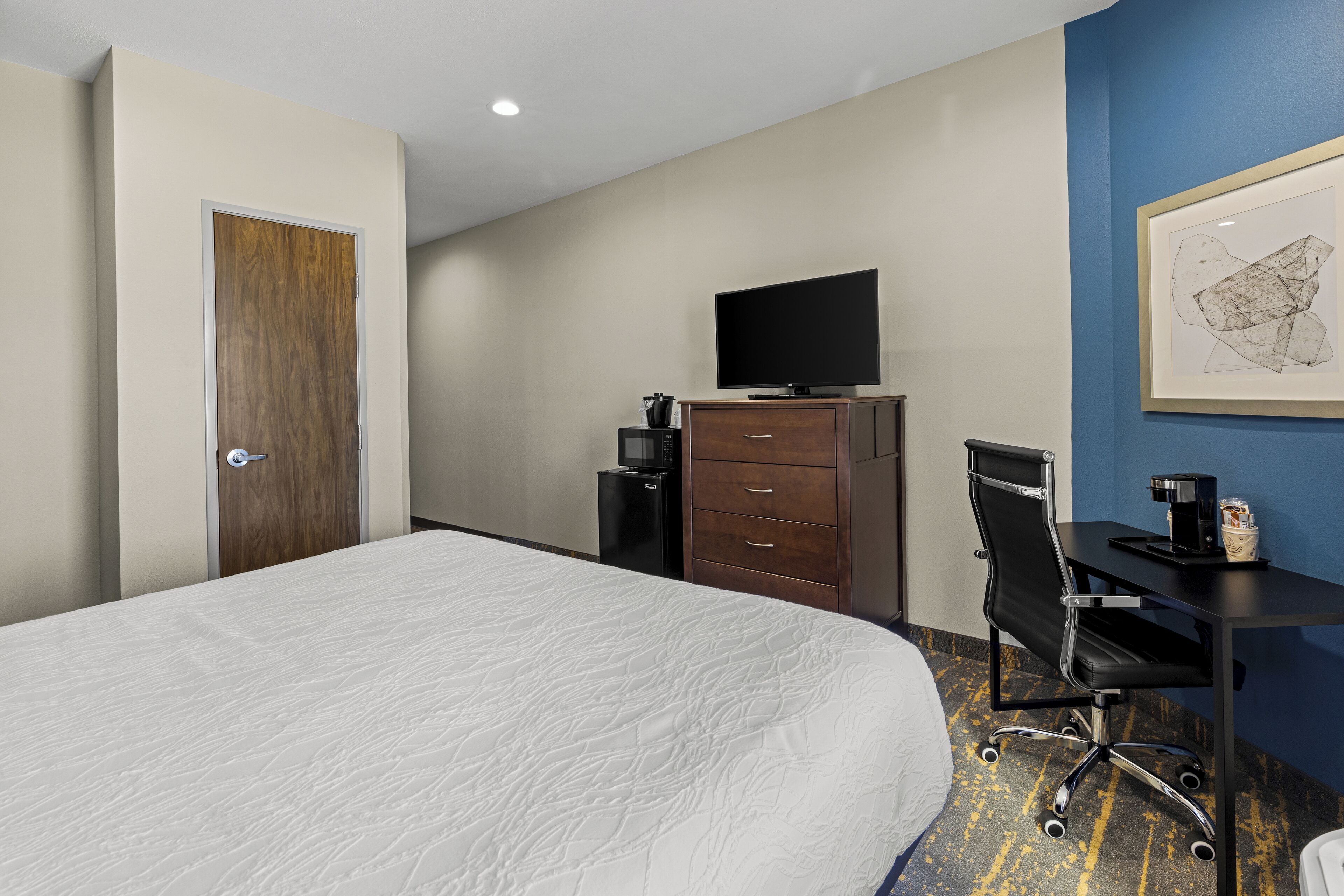 Habitación de hotel con una cama grande, un escritorio con silla, televisión, cafetera y una obra de arte enmarcada en la pared.