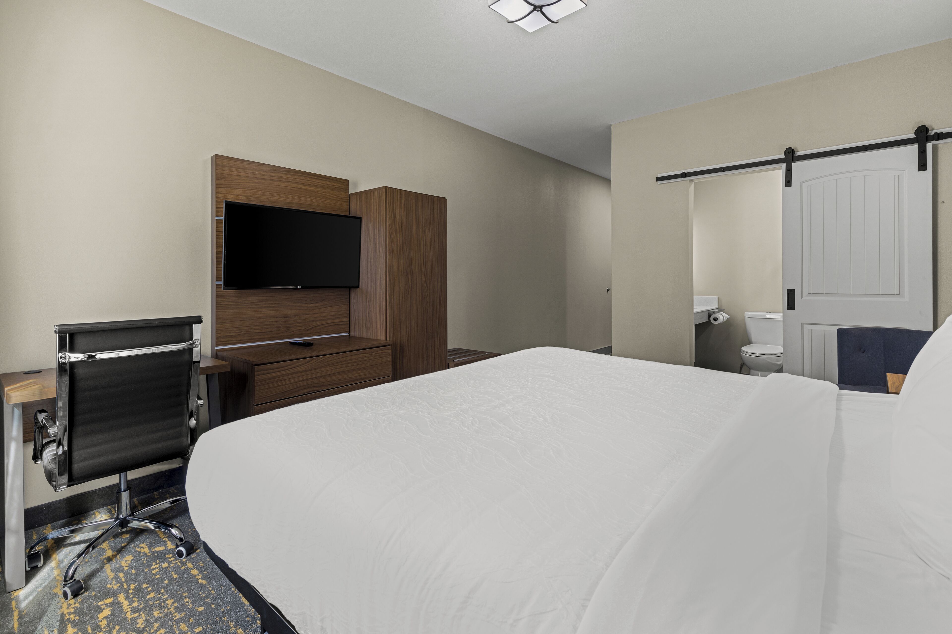 Une chambre d’hôtel comprenant un lit, un bureau avec un ordinateur, un téléviseur fixé au mur, et une salle de bain avec des toilettes et un lavabo.