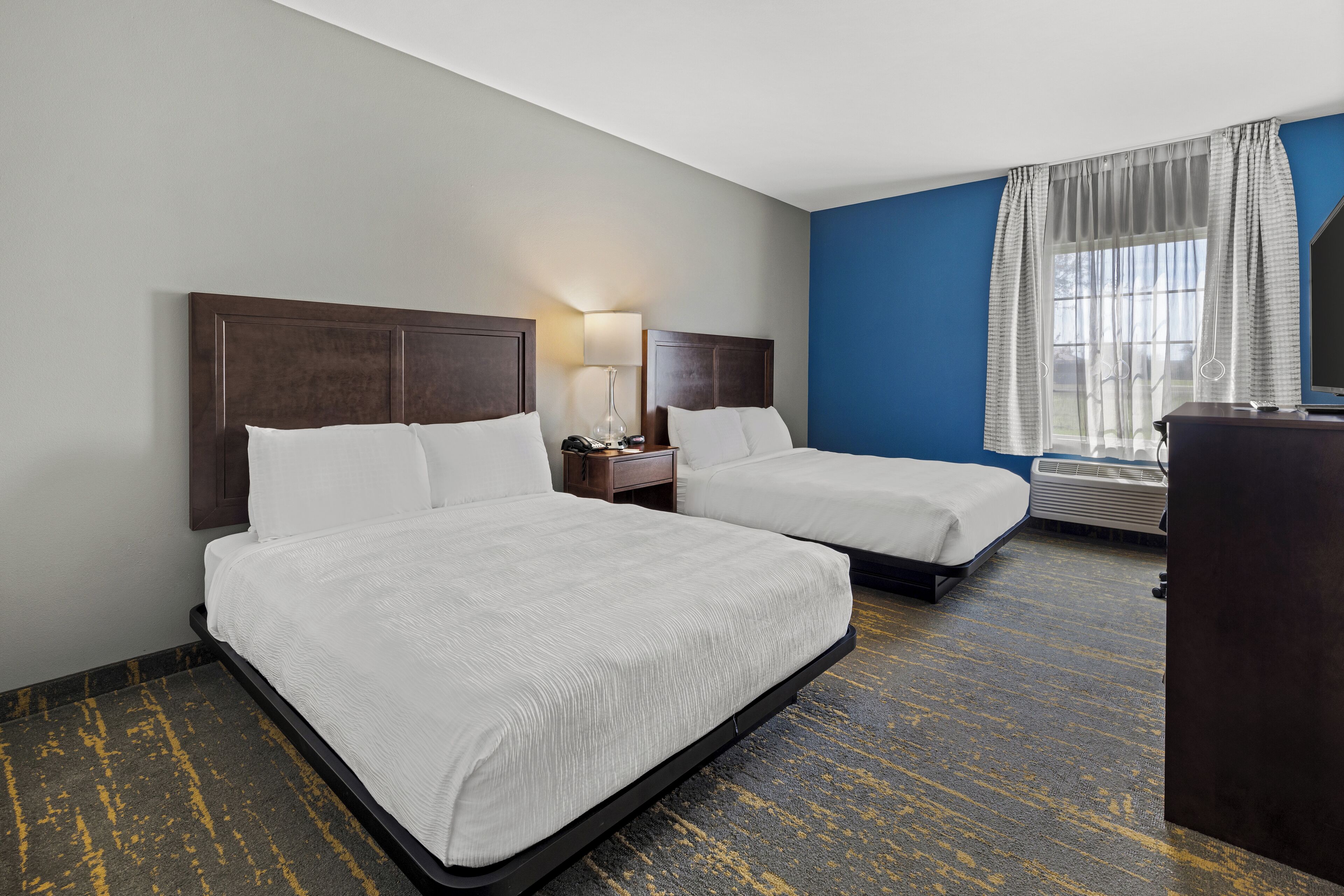 Ein Hotelzimmer mit zwei Betten, einem Schreibtisch, einer Lampe und einem großen Fenster mit Vorhängen.