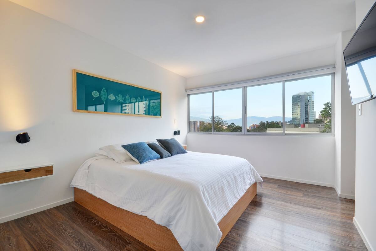A modern bedroom with a large bed, a view of the city, and a painting on the wall.