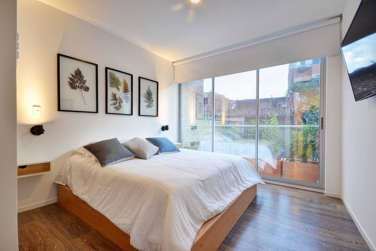A modern bedroom with a large bed, a nightstand, and framed botanical prints on the wall.
