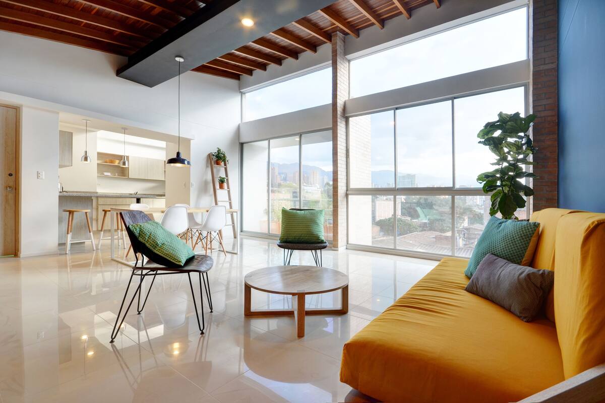 A spacious living area with a yellow sofa, a round wooden coffee table, and a large window offering a city view.
