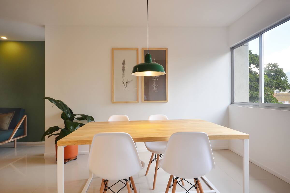 A modern dining area with a wooden table, white chairs, a green pendant light, and framed botanical prints on the wall.