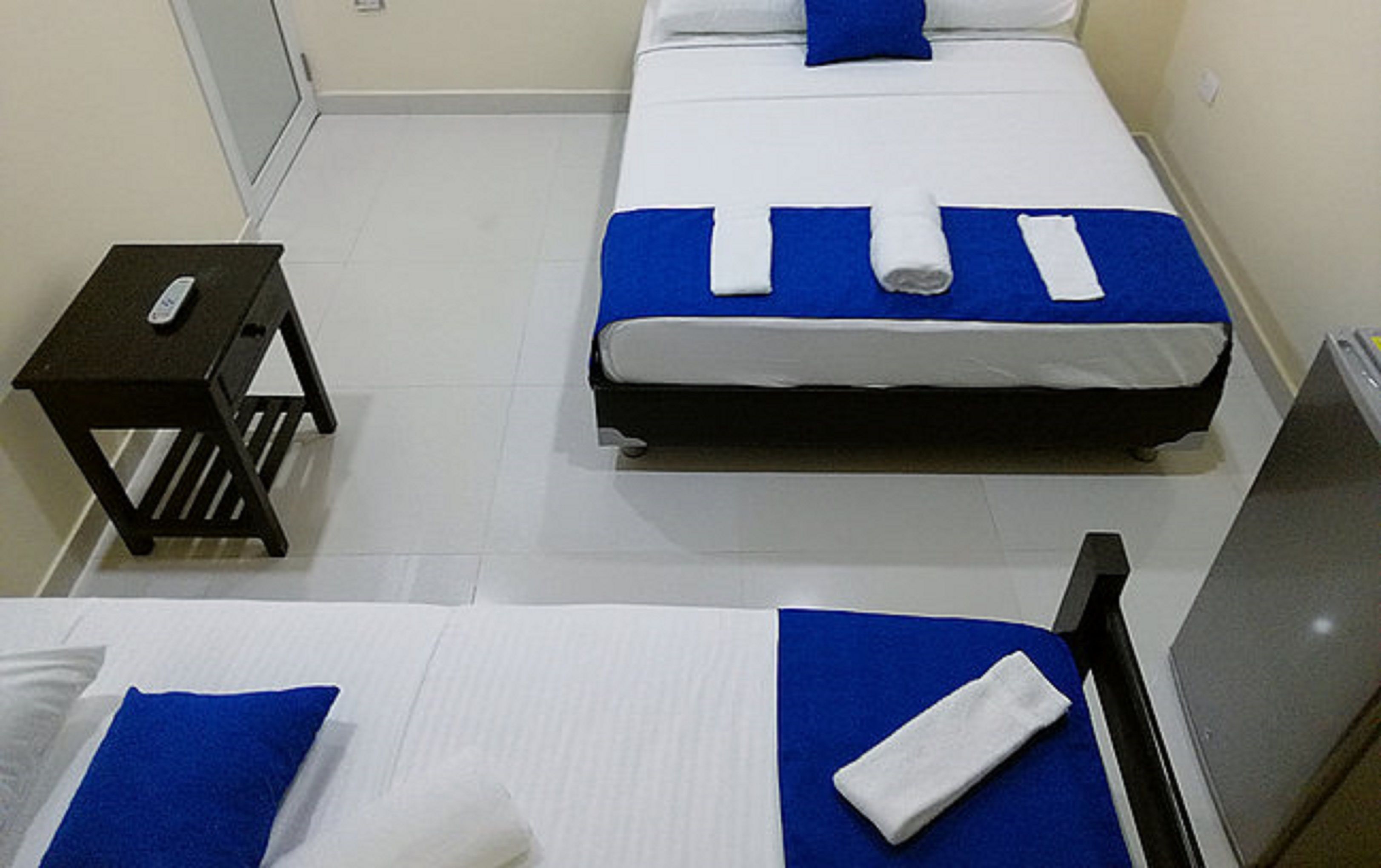 A hotel room with two beds, each with a blue bedspread and white pillows. There is a small black table with a remote control on it.