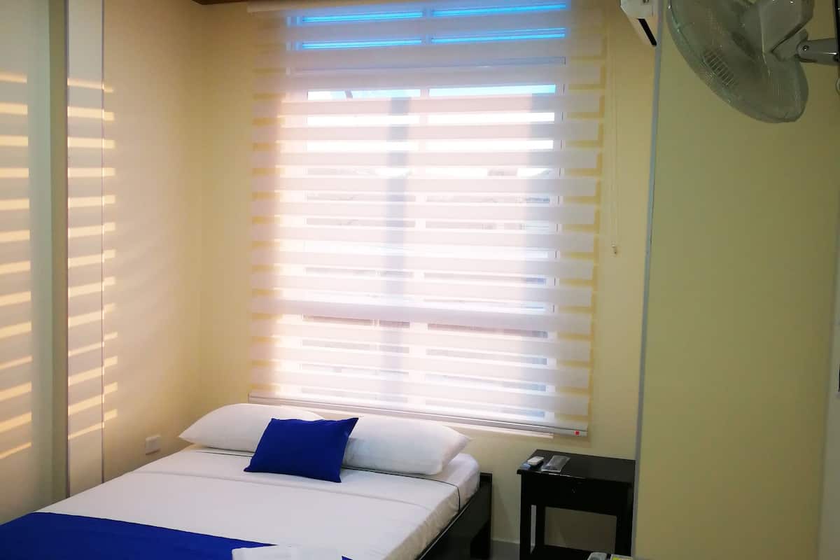 A bedroom with a bed, a bedside table, a fan, and a window with blinds.