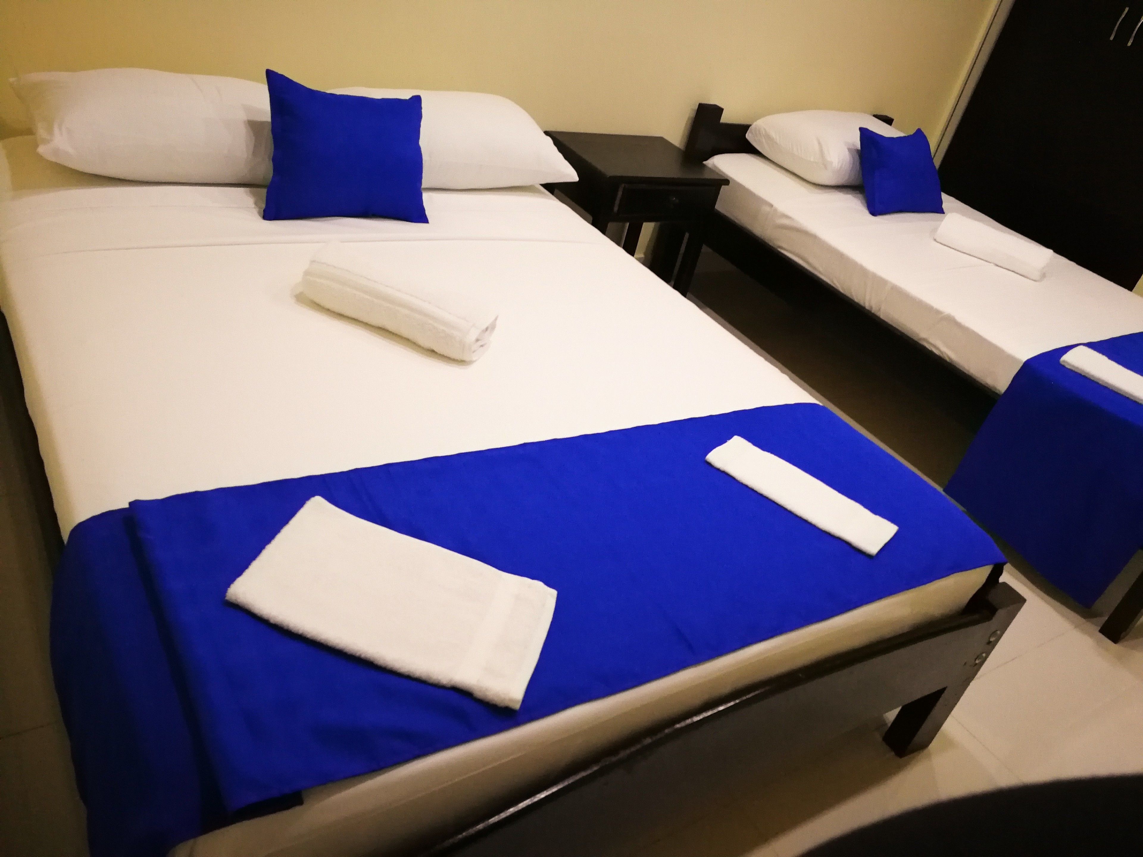 A hotel room with two single beds, each with a blue bedspread and white pillows. There is a small table between the beds and a black nightstand on the right.