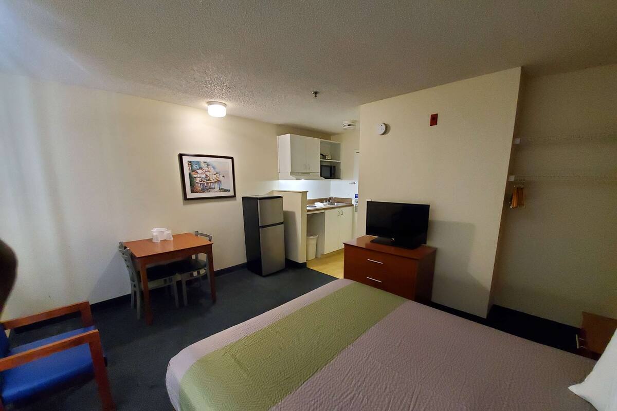 A hotel room with a bed, a desk with a chair, a refrigerator, a television, and a kitchenette.