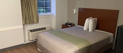 A hotel room with a large bed, a nightstand with a phone, a window with curtains, and a wall-mounted heater.