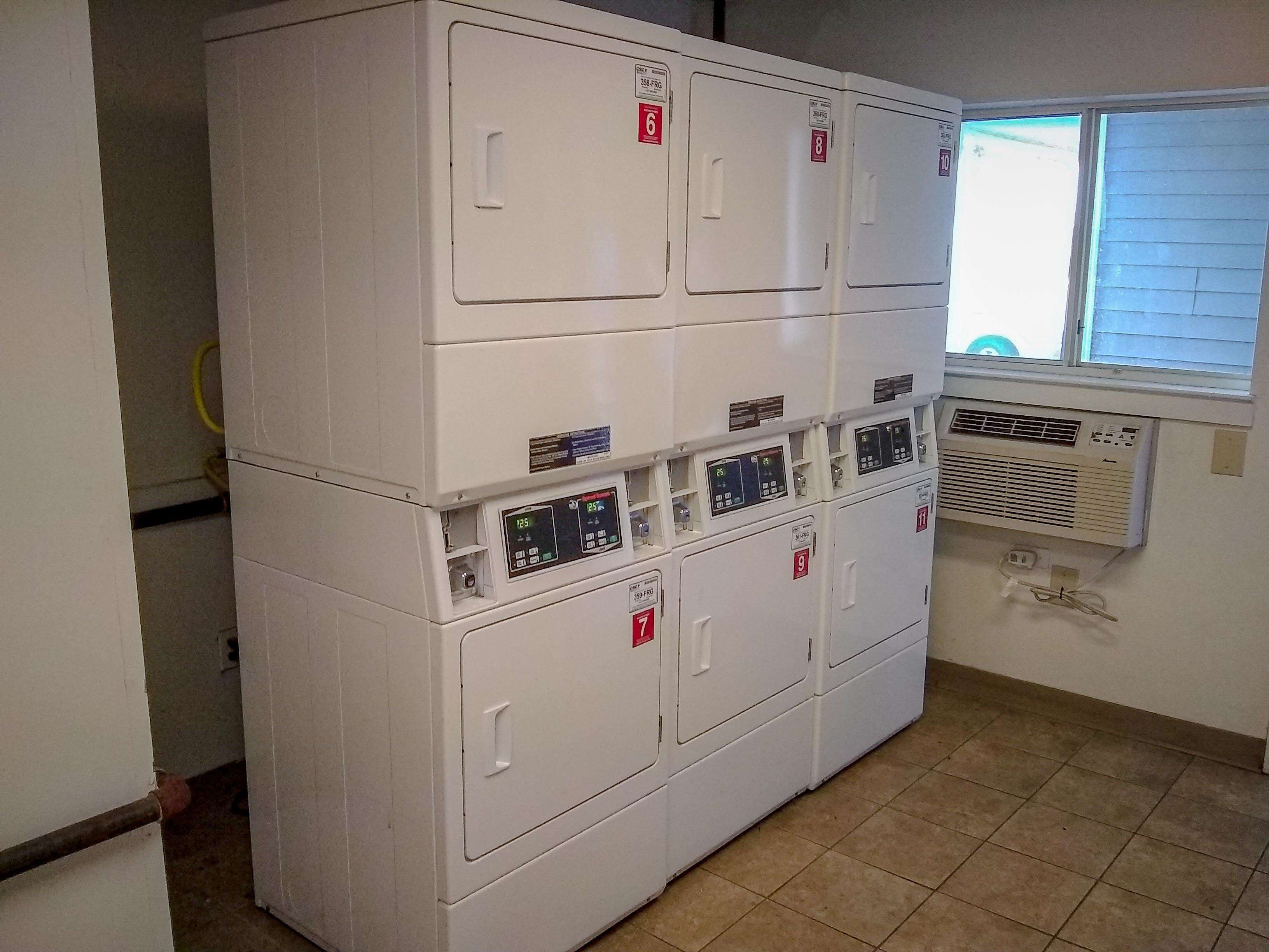 Een wasserette met gestapelde wasmachines en drogers, genummerd van 6 tot 11, en een airconditioningunit aan de muur bevestigd.