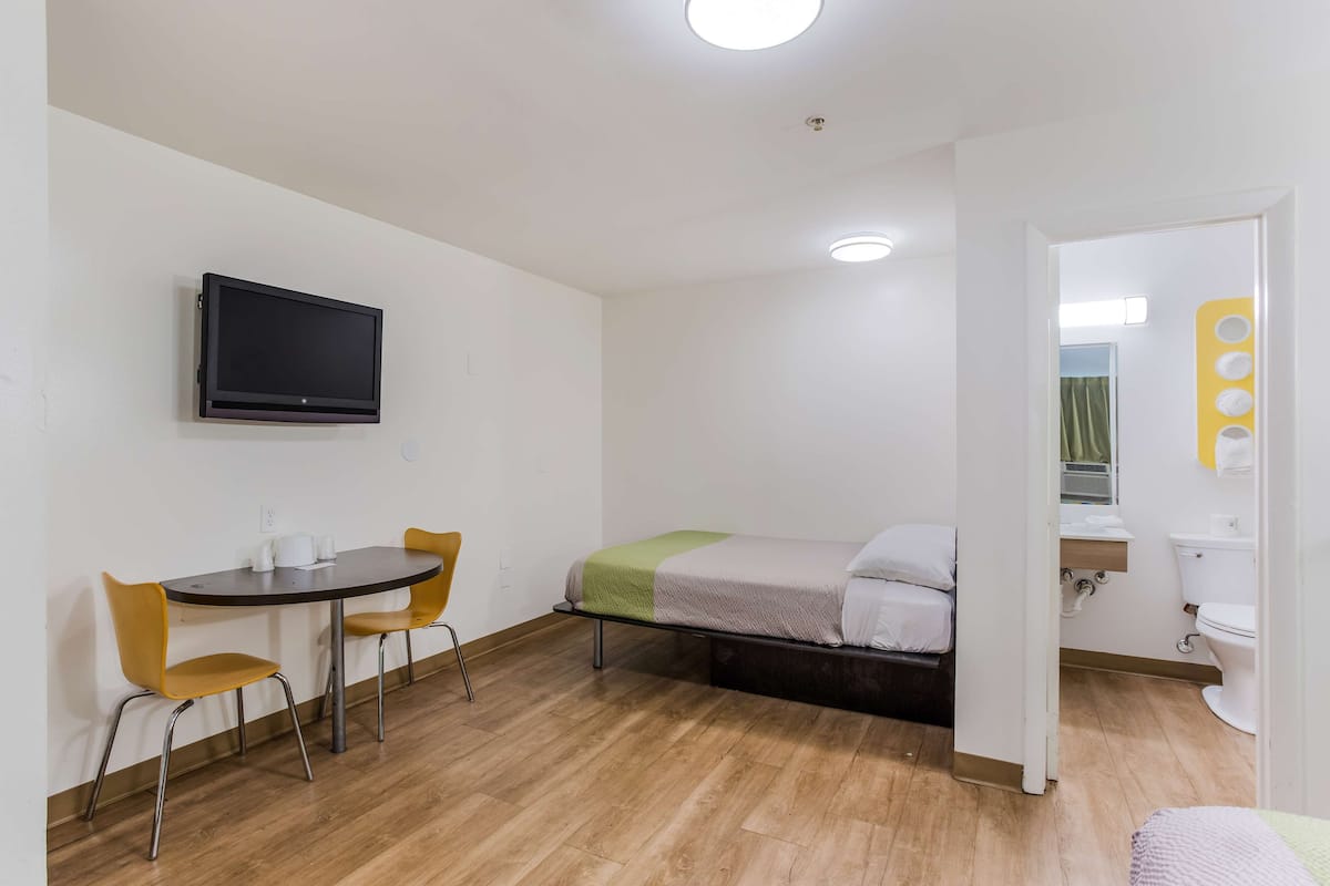 Una habitación pequeña y limpia con una cama, una mesa con sillas, un televisor montado en la pared y un baño visible a través de una puerta abierta.