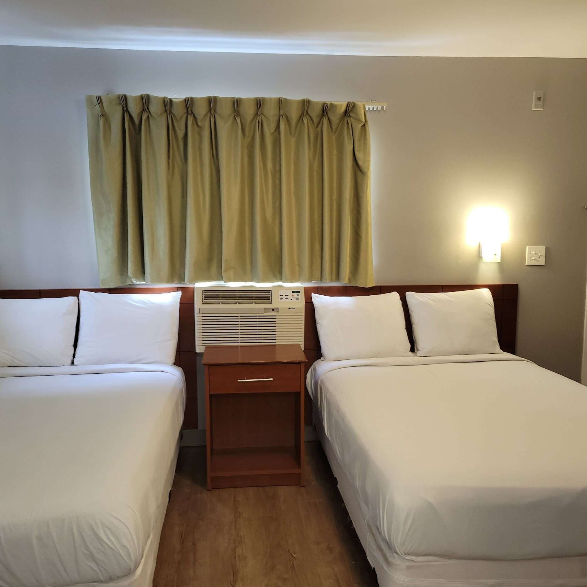 A hotel room with two single beds, a nightstand, an air conditioning unit, a window with curtains, and a wall-mounted light.