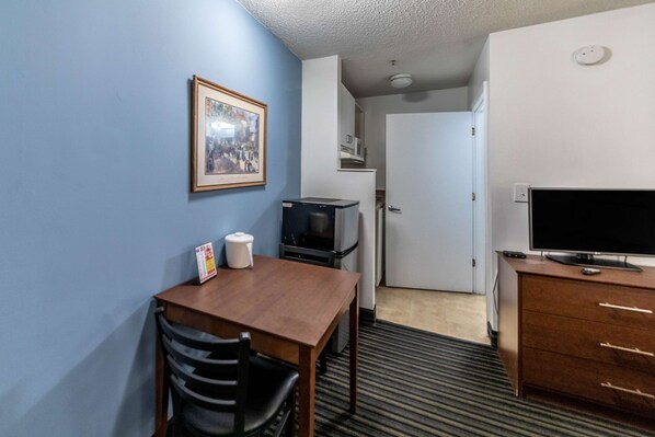 A compact hotel room with a wooden dining table, a TV on a dresser, a framed picture on the wall, and a microwave.
