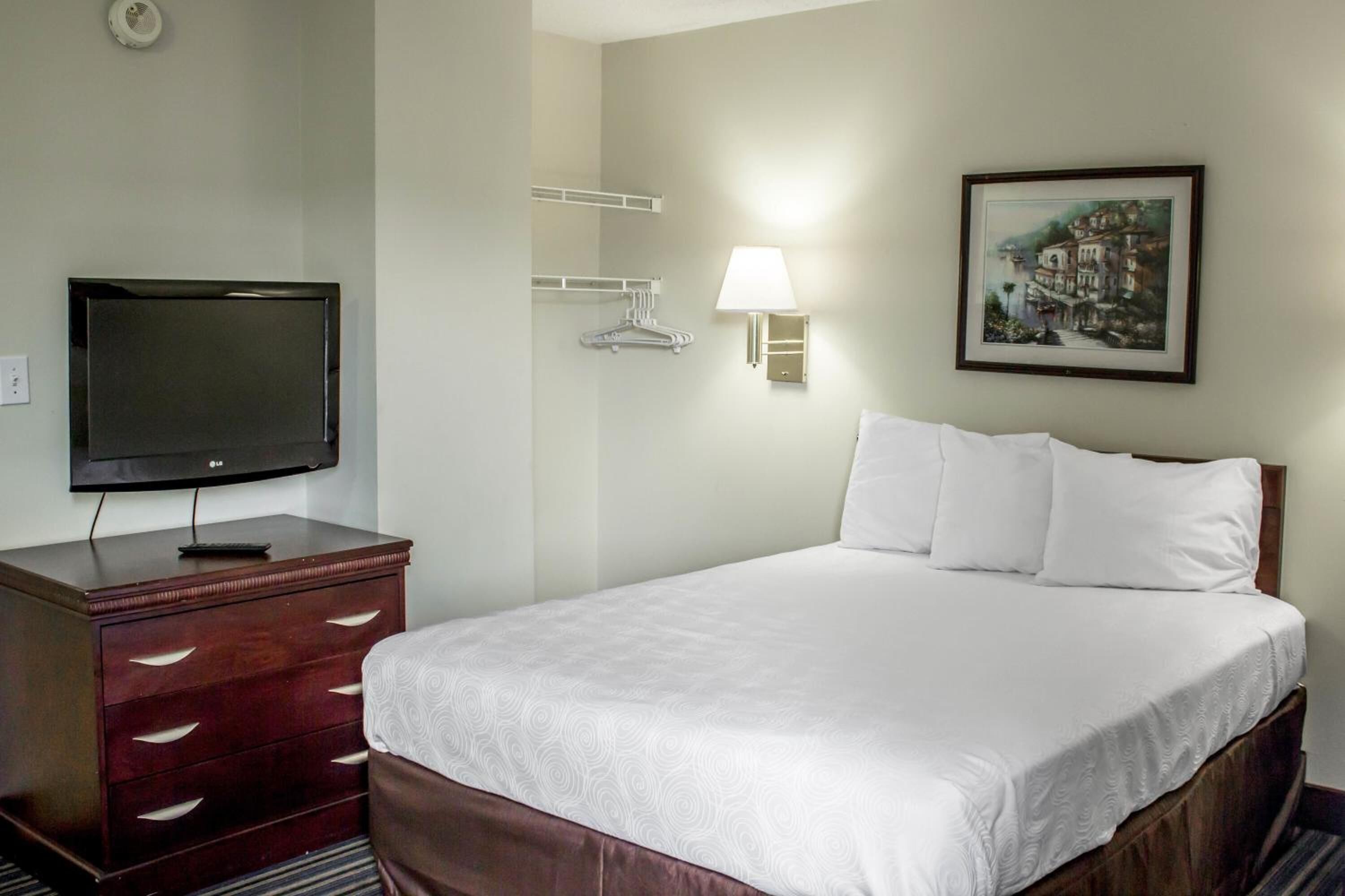 Habitación de hotel con una cama, un televisor sobre una cómoda de madera, una lámpara de pared y un cuadro enmarcado.