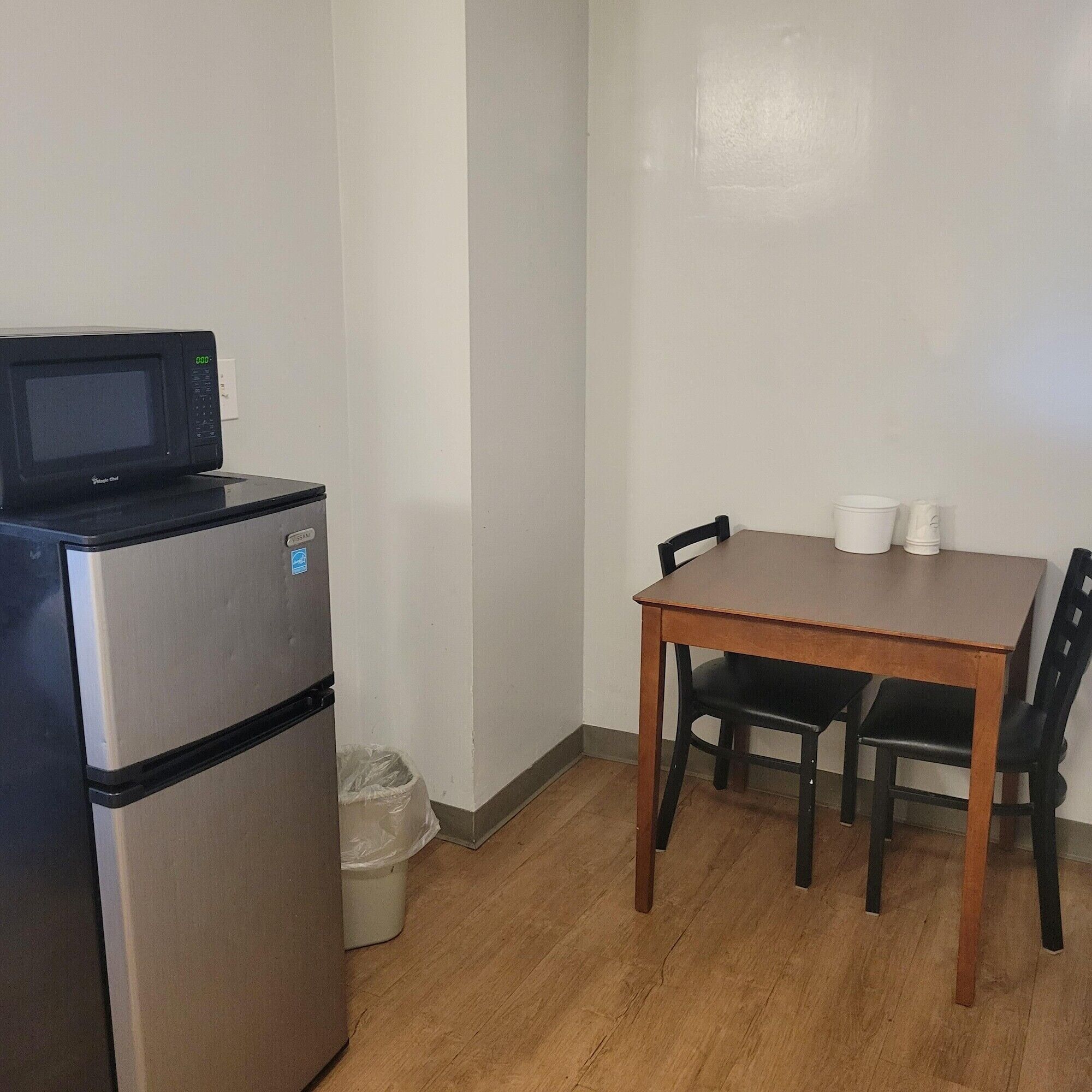 A compact kitchenette with a microwave and refrigerator, a wooden dining table with four chairs, and a trash bin.