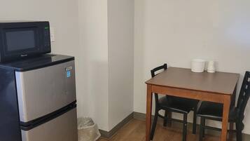 A compact kitchenette with a microwave and refrigerator, a wooden dining table with four chairs, and a trash bin.
