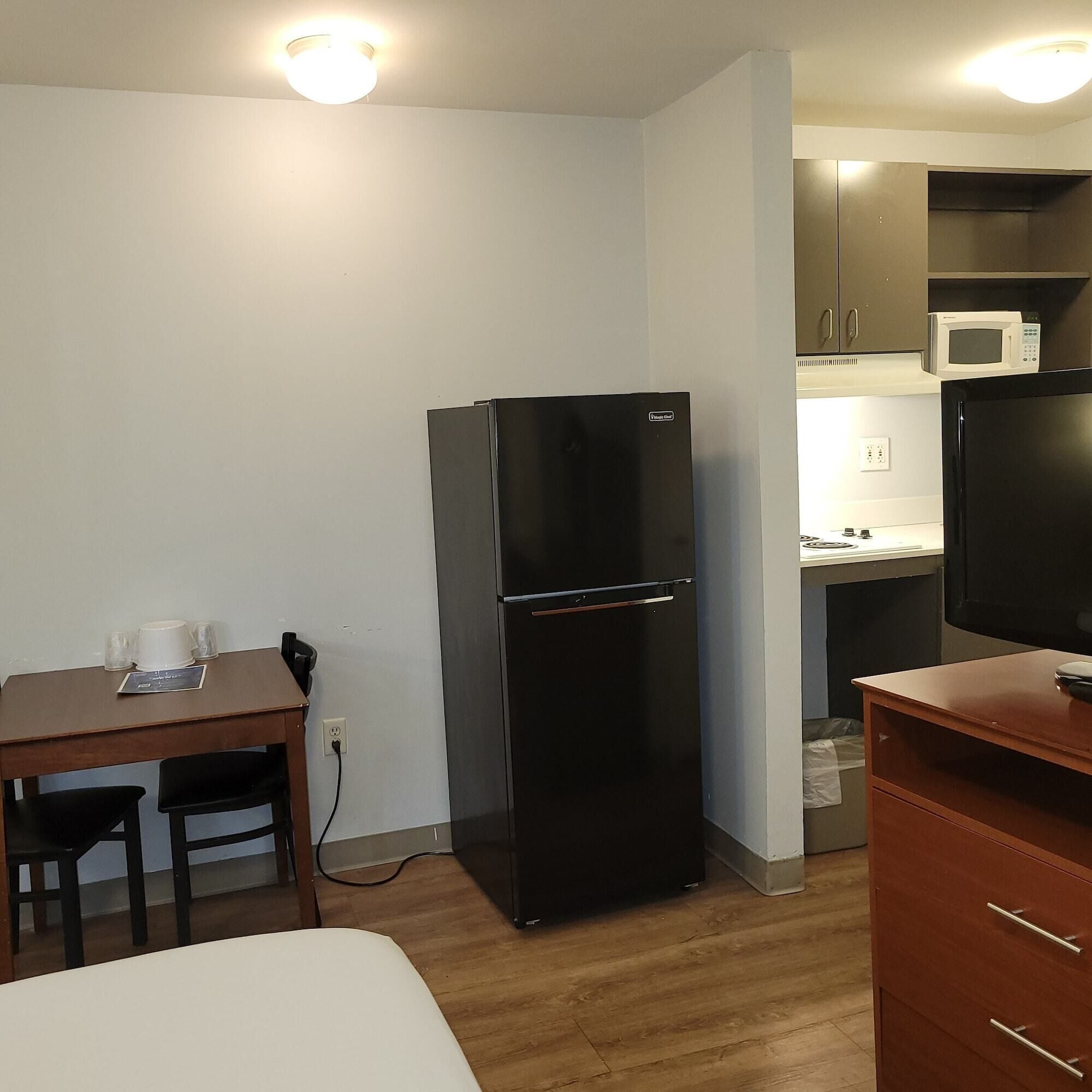 A compact kitchenette with a black refrigerator, a microwave, and a sink. There's a small dining table with chairs and a single bed in the room.