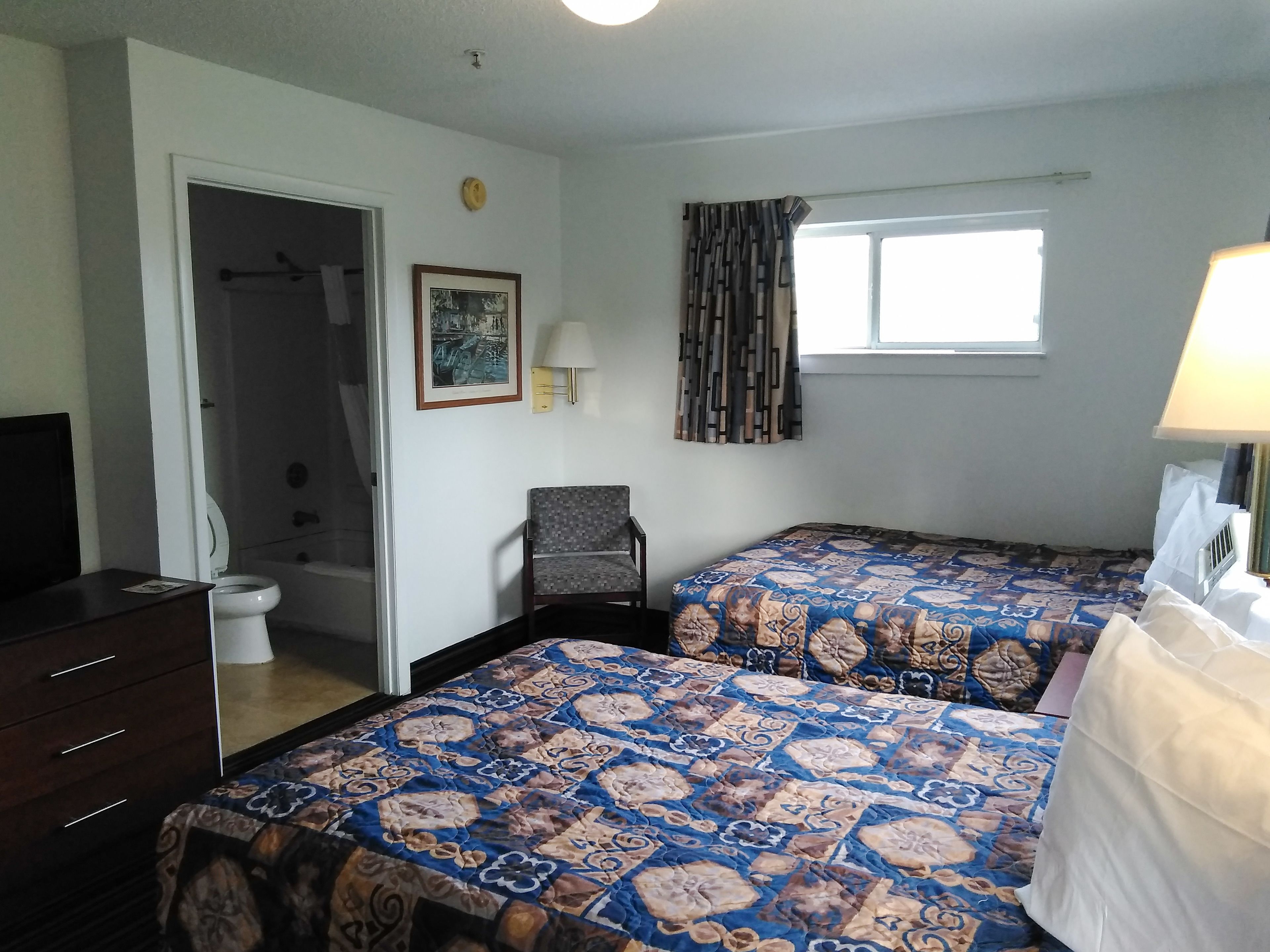 A bedroom with two beds, a chair, a desk, a window with curtains, a wall clock, and a framed picture.