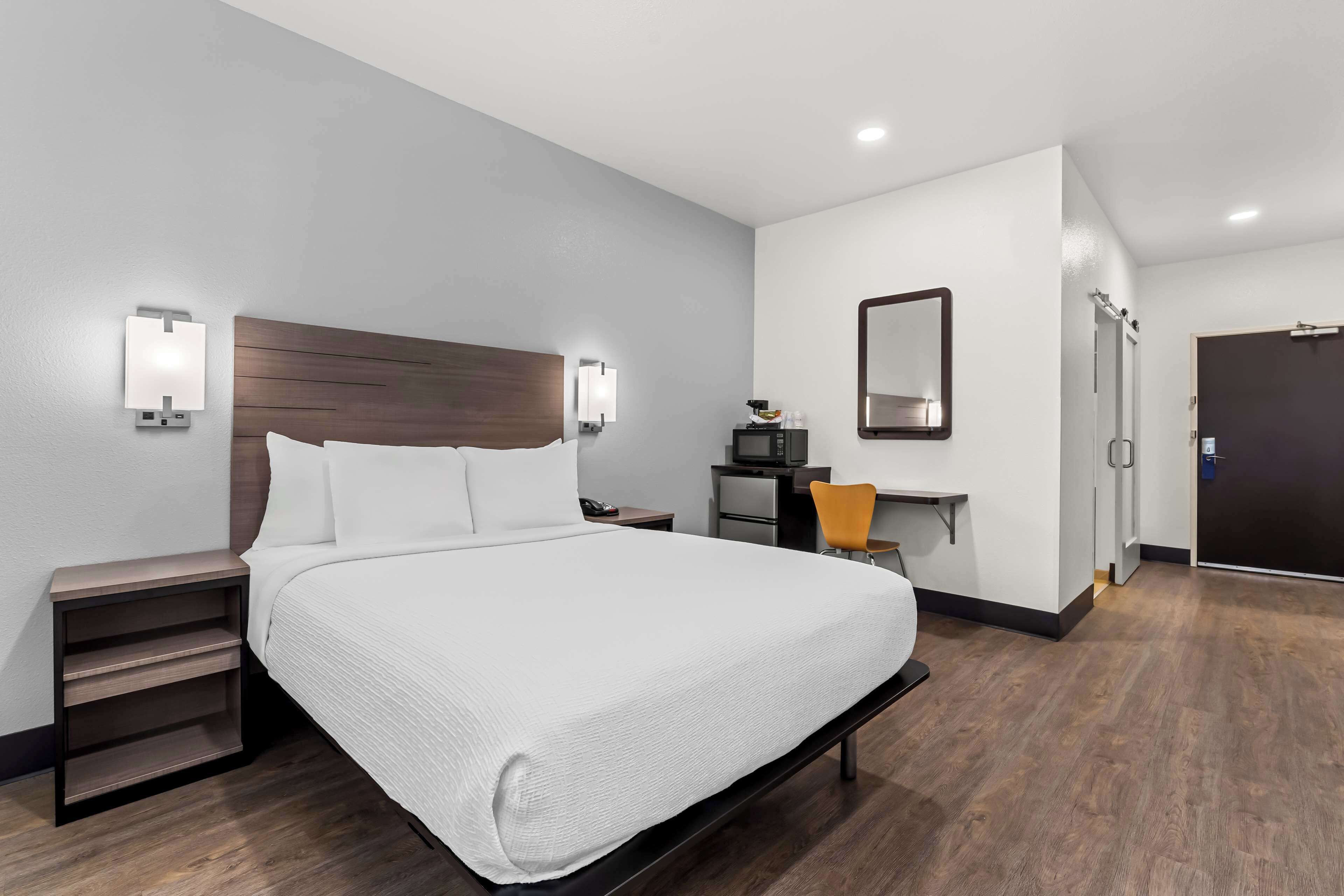 A modern hotel room with a large bed, bedside tables, a desk with a chair, a TV, and a door.