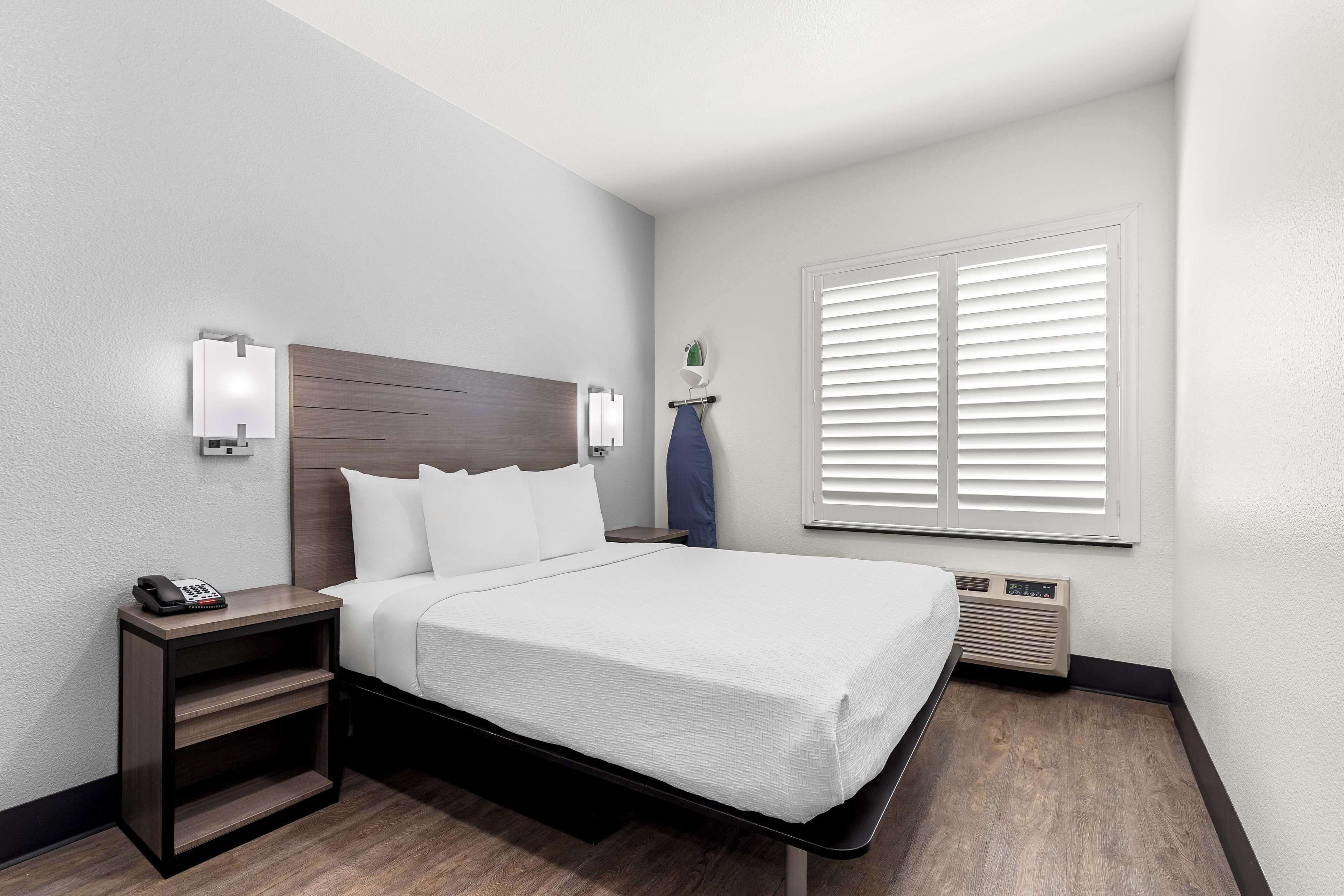 A hotel room with a bed, a nightstand with a phone, a wall-mounted light, a window with blinds, and a wall-mounted heater.