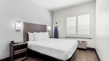 A hotel room with a bed, a nightstand with a phone, a wall-mounted light, a window with blinds, and a wall-mounted heater.