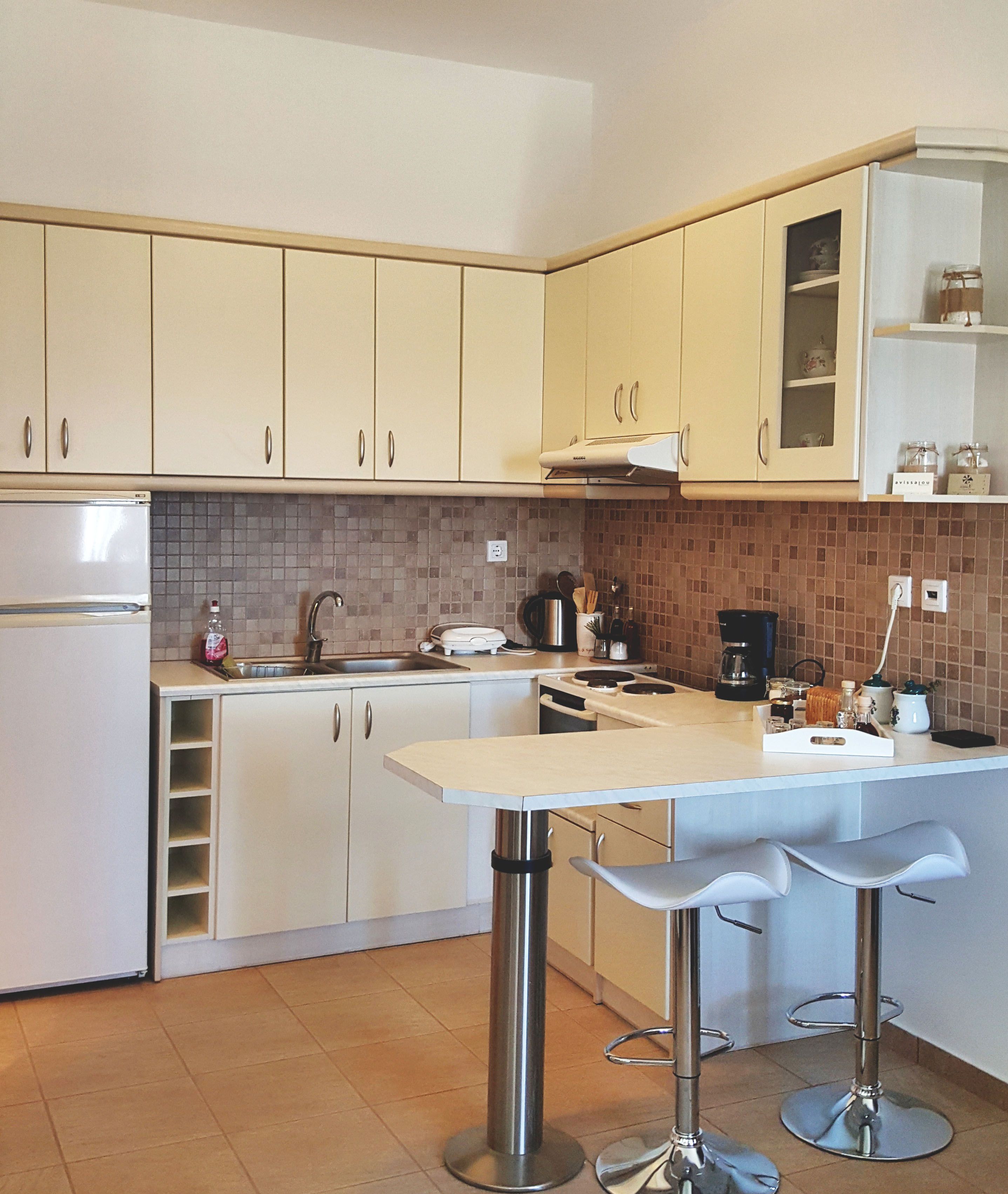 A modern kitchen with white cabinets, a stainless steel refrigerator, a double sink, and a bar area with stools.
