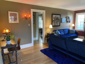 Un salon avec un canapé bleu, une petite table avec une lampe et un tapis bleu. Une porte donne accès à une autre pièce.