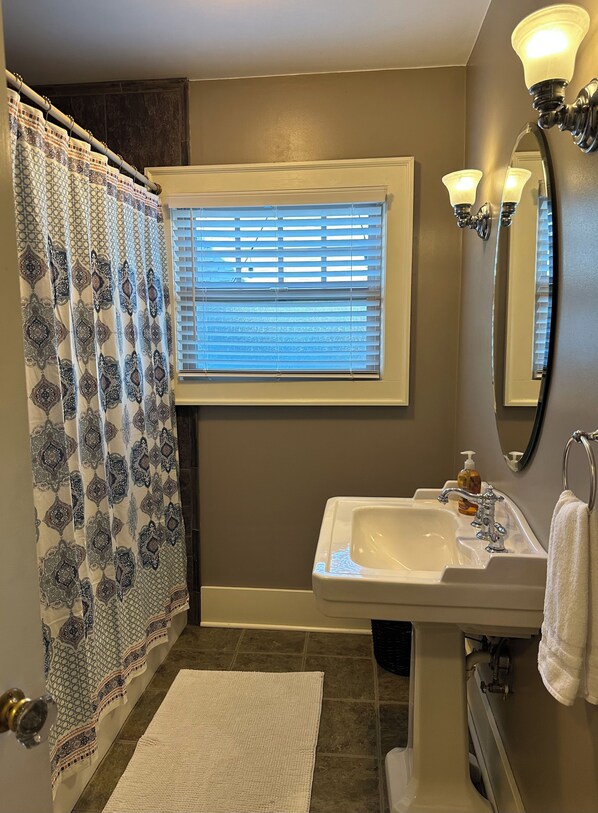 A bathroom with a shower curtain, a window with blinds, a sink with a mirror, and a towel rack.