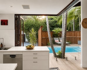 A modern kitchen with a view of a backyard swimming pool, wooden fence, and tropical plants. - Luxe Beachfront Haven w/heated pool. Pet-Friendly (Broken Head)