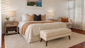 Ein Schlafzimmer mit Bett, Nachttischen, einer Bank, einem Gemälde und einem Fenster mit Vorhängen.