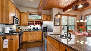 A kitchen with wooden cabinets, stainless steel appliances, a granite countertop, and a dining area with a red tablecloth. - Scenic Luxury Home - Hollows Resort & Marina, Lake Travis, 4 Pools (Jonestown)