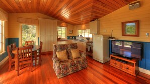 A cozy cabin interior with a wooden floor, a floral sofa, a dining table with chairs, a kitchenette, and a television. - Island View Spa Cottage (Smithton)