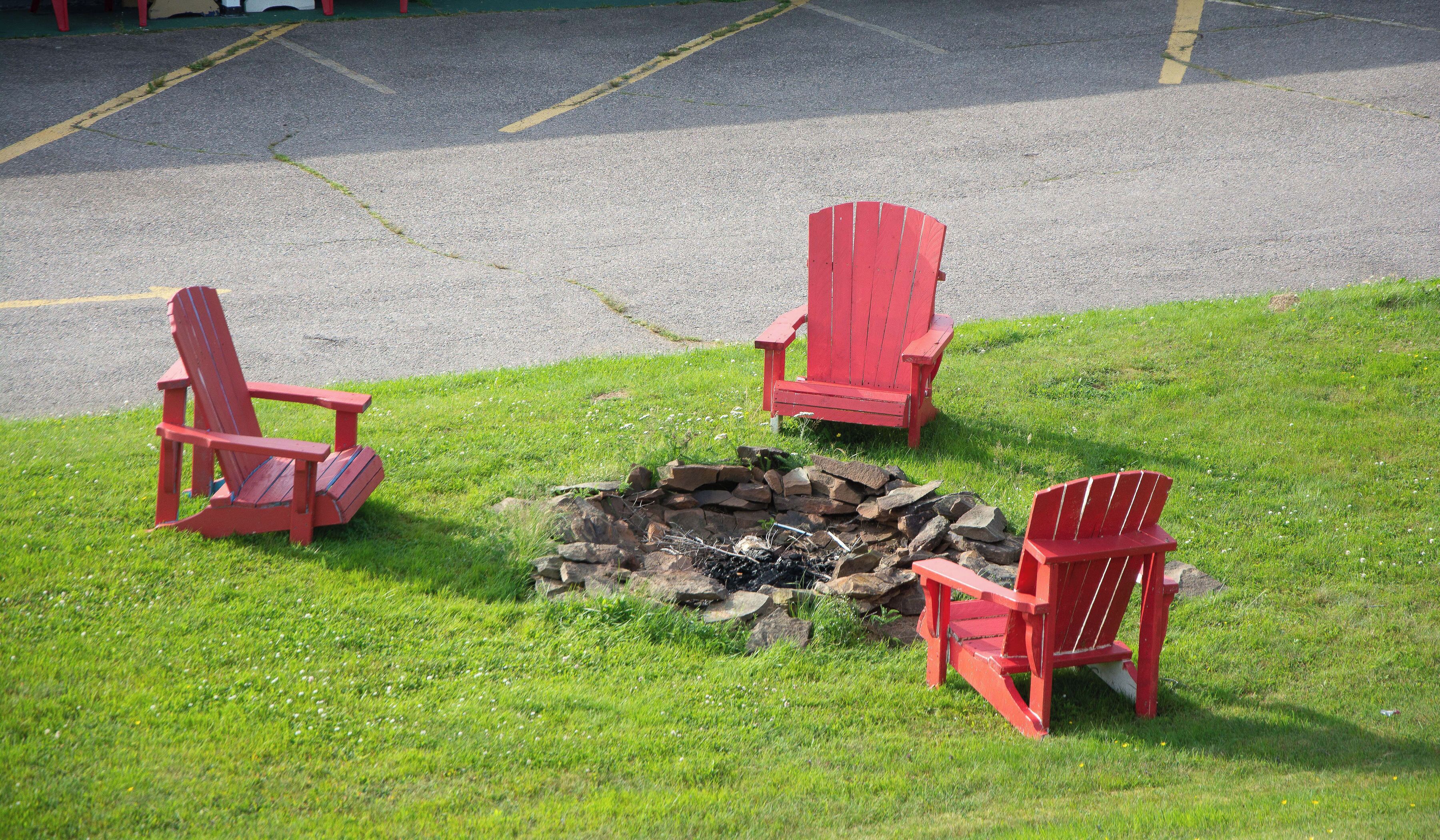 Three red Adirondack chairs arranged around a stone fire pit on a grassy area adjacent to a paved road.