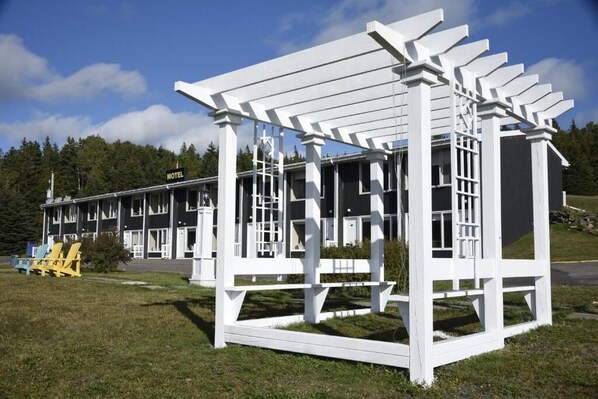 Una estructura blanca de madera con diseño de pérgola, un motel al fondo y un área verde con sillas amarillas.