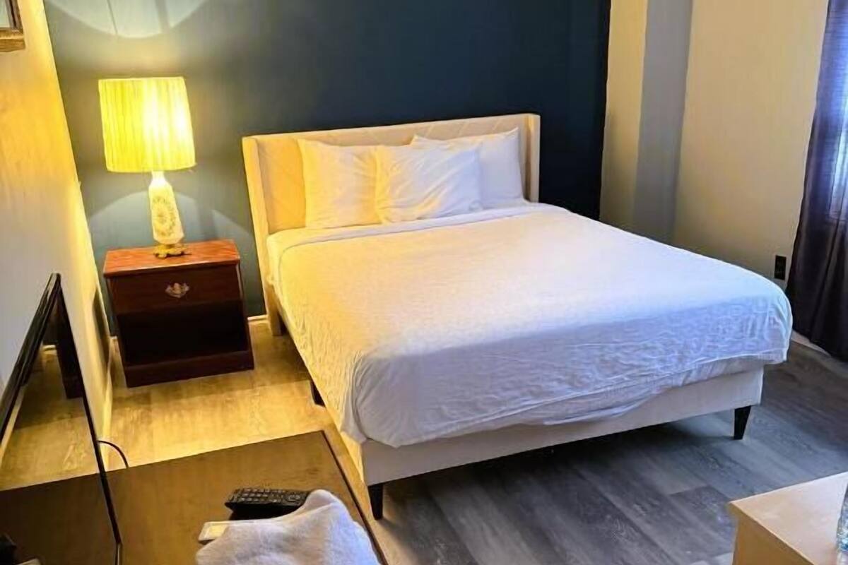 Een hotelkamer met een bed, nachtkastje, lamp en een afstandsbediening op de vloer.